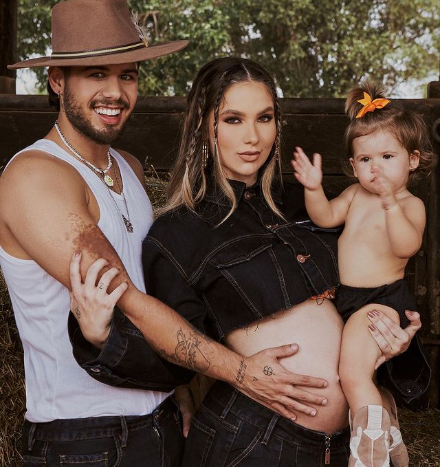 Zé Felipe acaricia a barriga de Virginia Fonseca grávida, ela segura sua filha Maria Alice no colo. Todos sorriem para a câmera