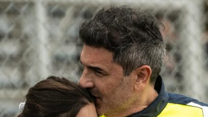 Homem beija a testa de uma mulher em um evento esportivo, com uma grade ao fundo.