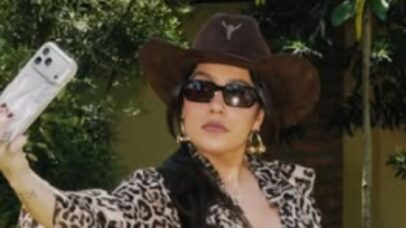 Mulher com chapéu de cowboy e óculos escuros tira selfie com celular, vestindo jaqueta estampada de onça.