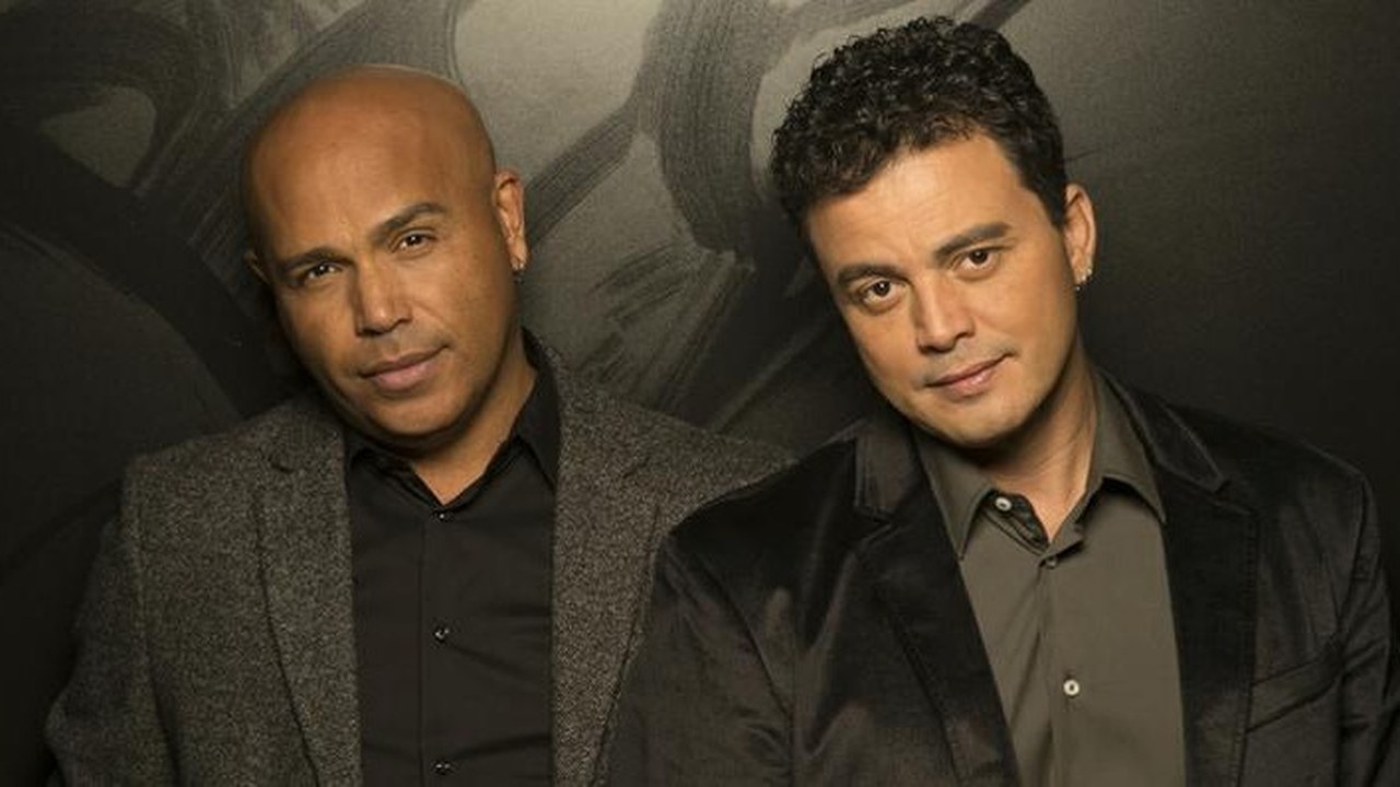 Rick e Renner, os dois cantores, posam juntos de casaco em um fundo escuro