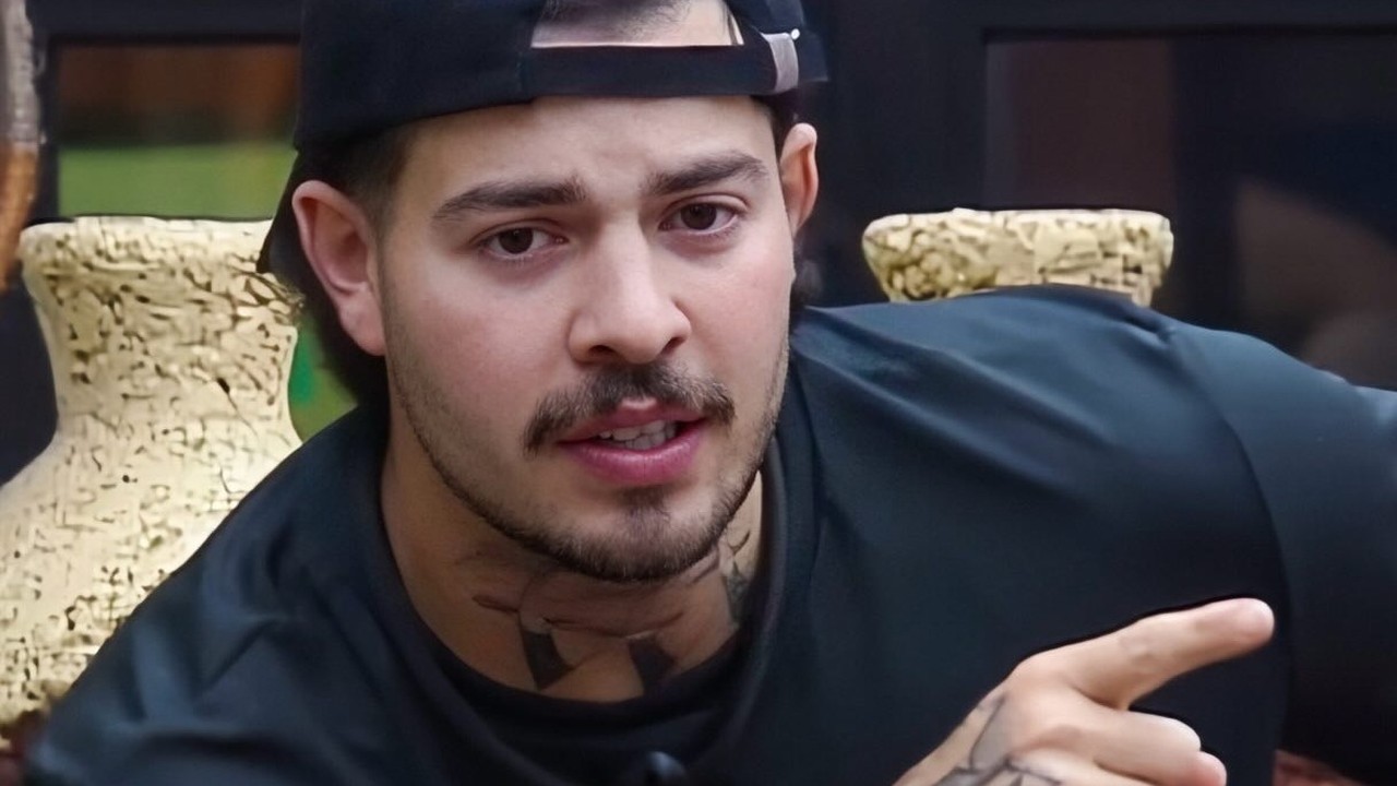 Homem, camisa preta e boné, com barba e tatuagens, aponta o dedo indicador