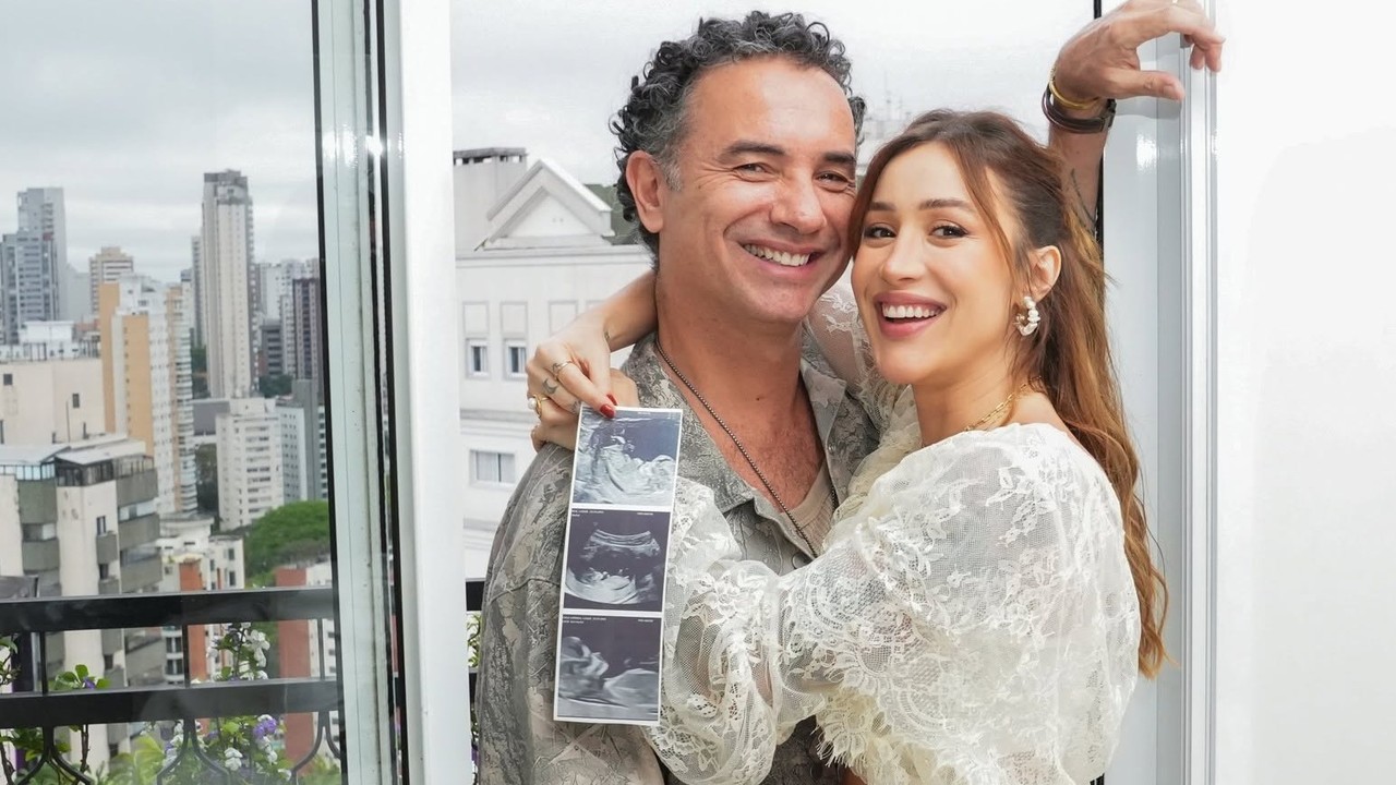Marco Luque e Jessica Correia sorriem, abraçados, segurando ultrassom do bebê, com vista da cidade