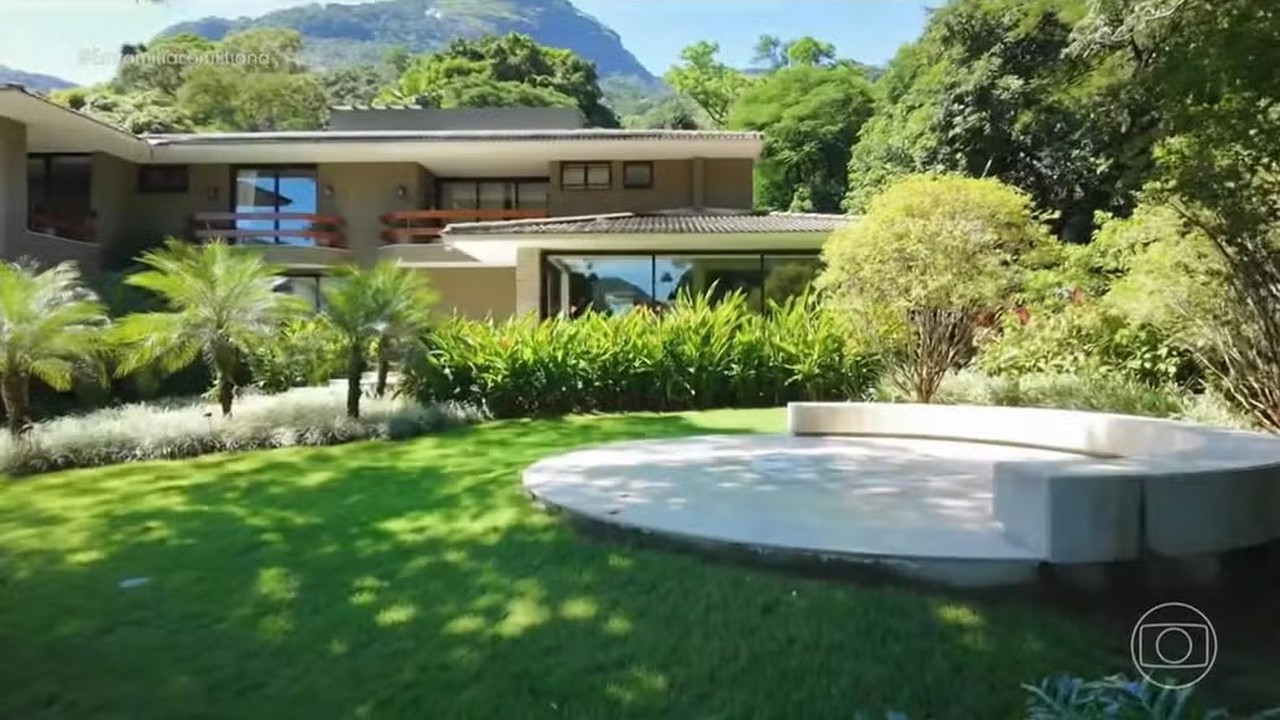 Casa moderna, jardim verde, palmeiras, pátio circular, montanhas e logo Em Família com Eliana