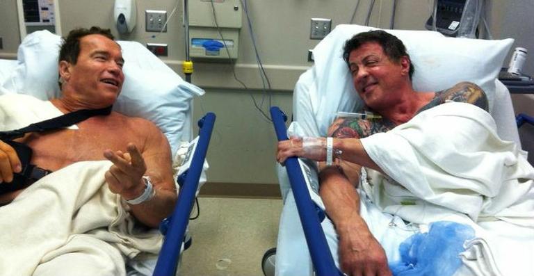 Arnold Schwarzenegger e Sylvester Stallone conversam em cama de hospital